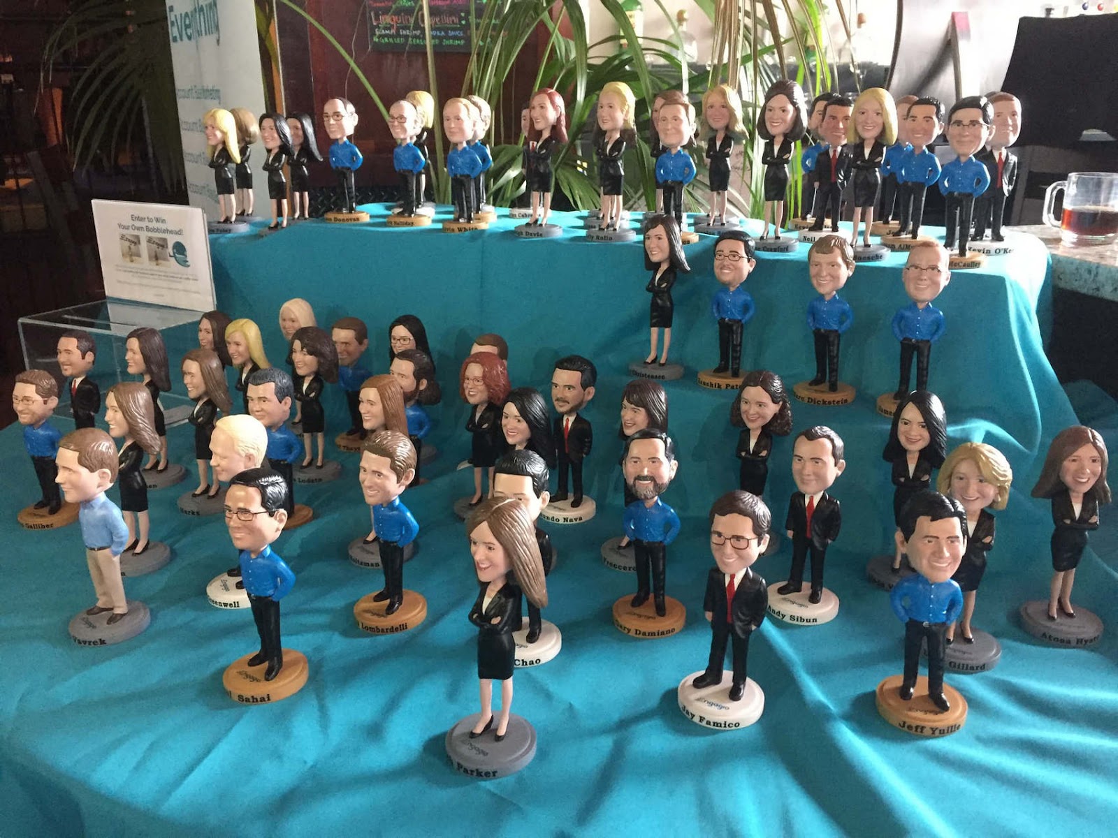 Custom bobblehead figurines displayed on a teal-covered table in rows, with some on raised platforms.