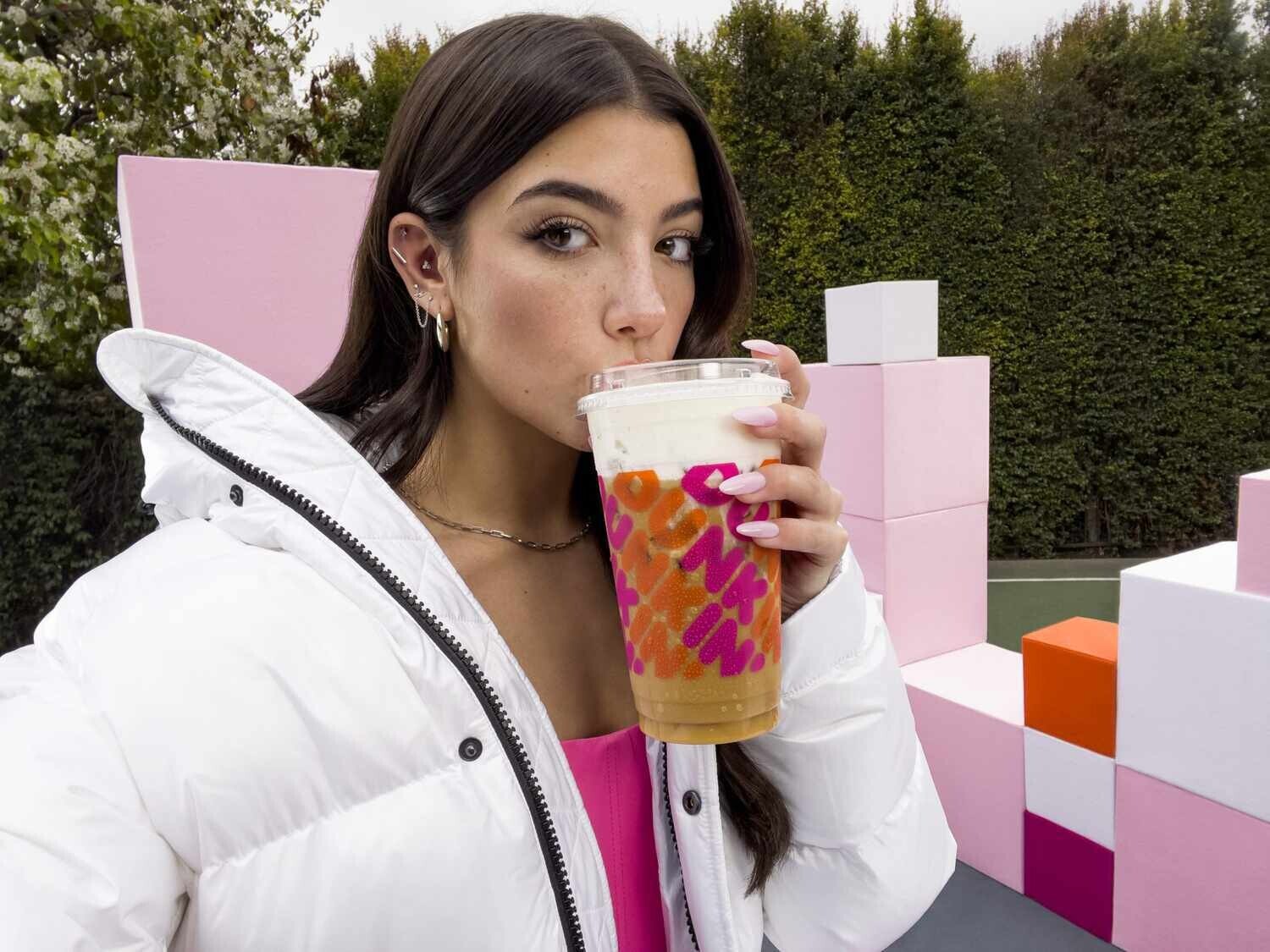 A young woman with long brown hair is pictured wearing a white winter coat and sipping from a large Dunkin’ Donuts iced coffee. 